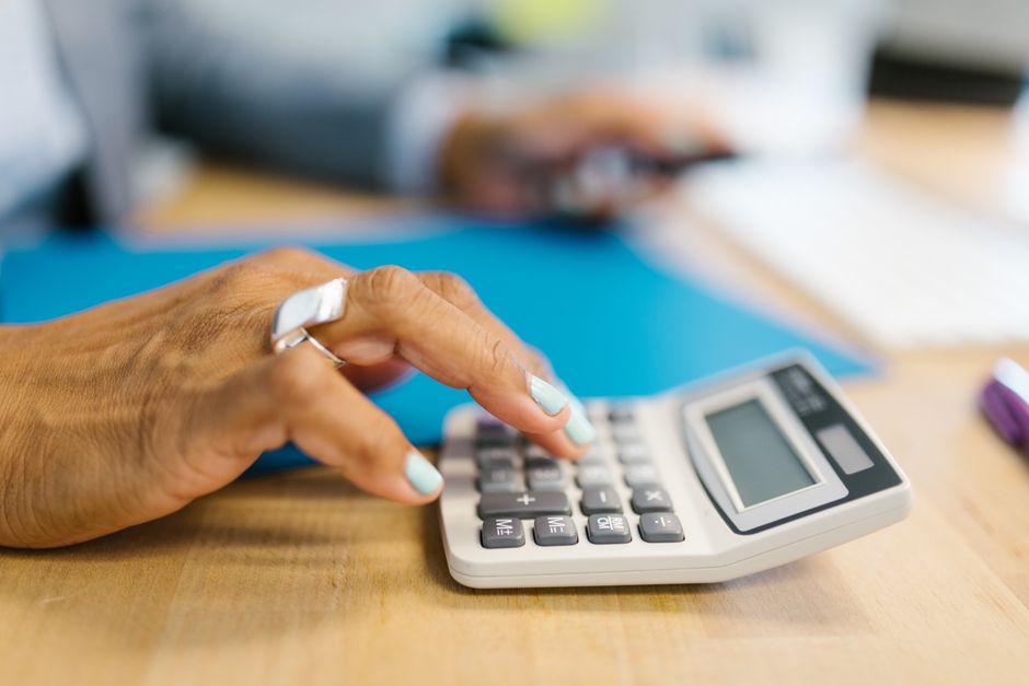 A person sits at a modern table with a calculator and documents, thoughtfully examining a small architectural model of a house, illustrating the process of making an informed financial decision about real estate commissions.
